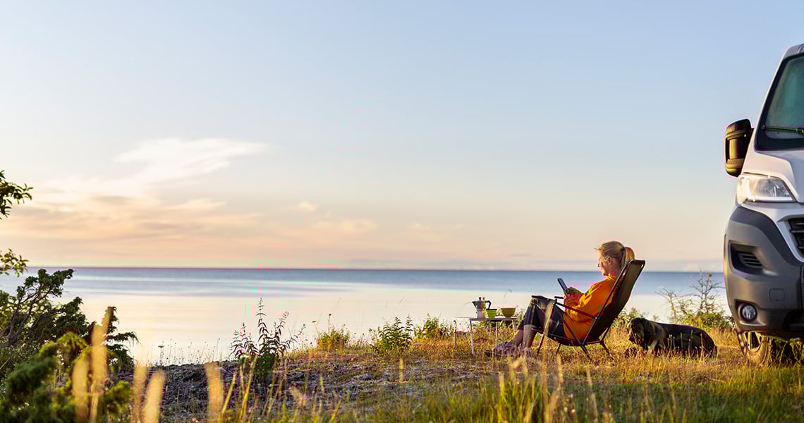 Kvinna med hund sitter vid havet i solstol bredvid husbil