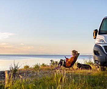 Kvinna med hund sitter vid havet i solstol bredvid husbil